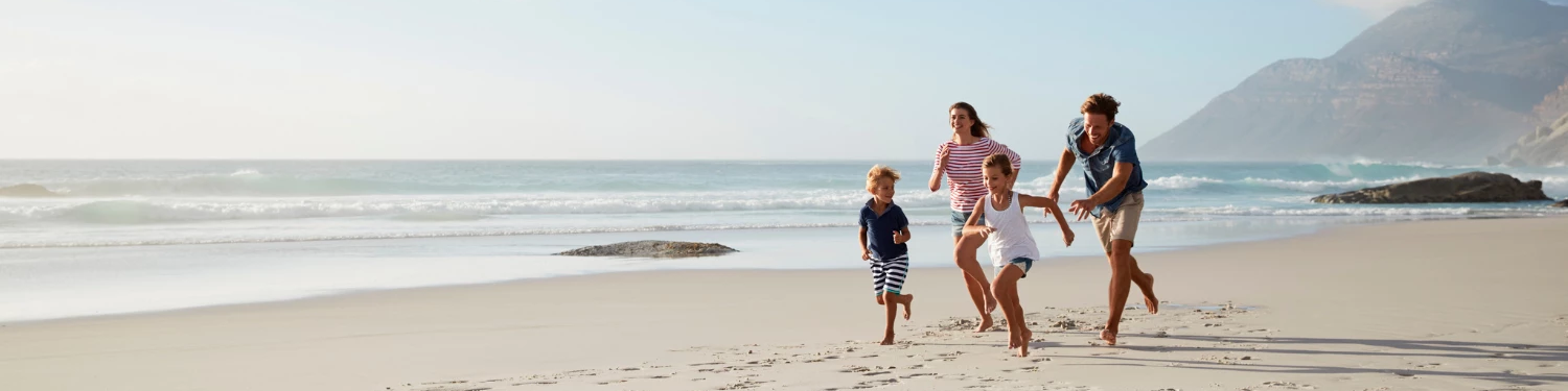 A family of four runs barefoot along a sunny beach with waves behind them and mountains in the distance, smiling together.