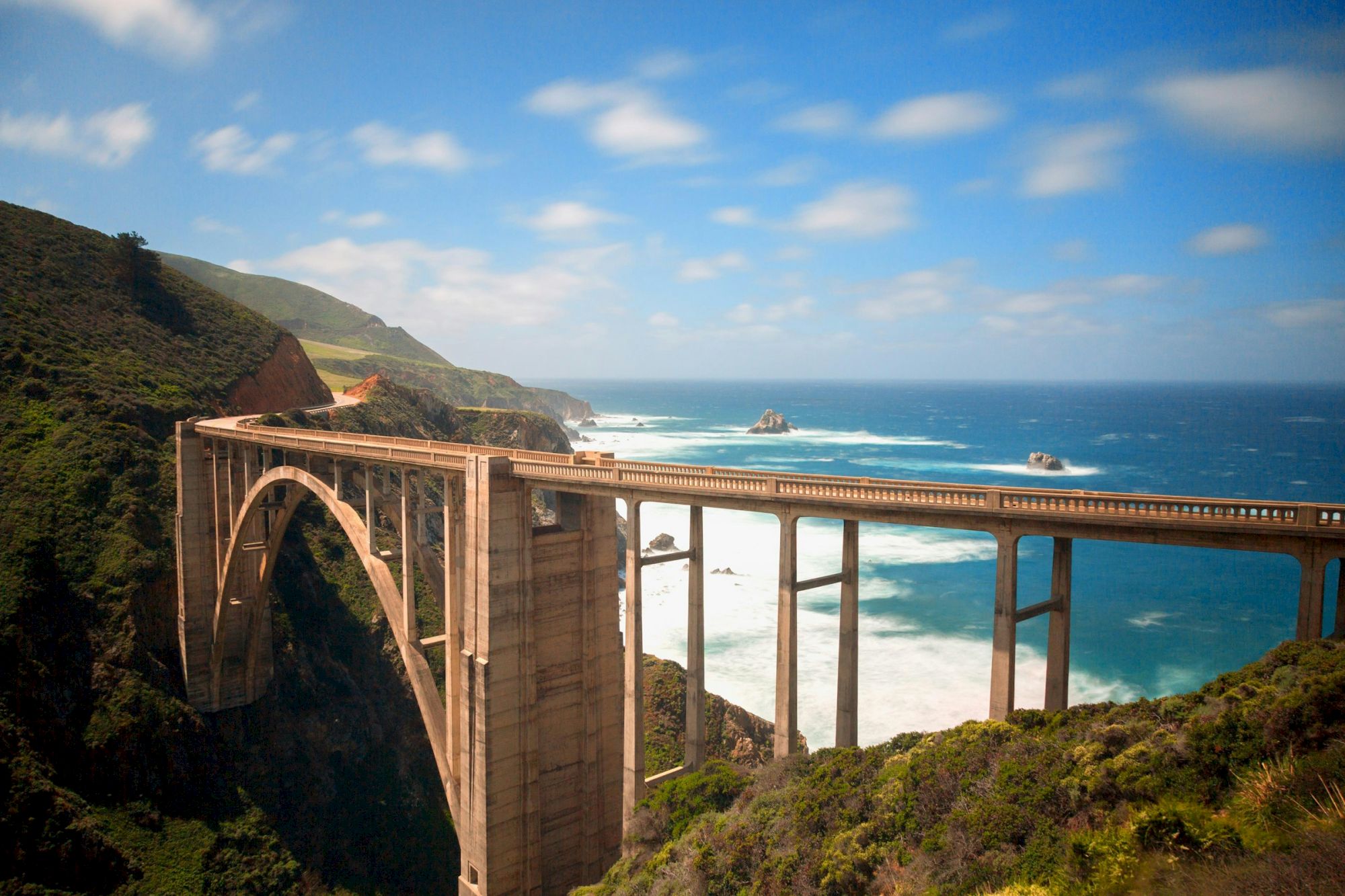 A highway viaduct arches along a rugged coastline, carrying cars above crashing waves and rocky islets under blue sky.