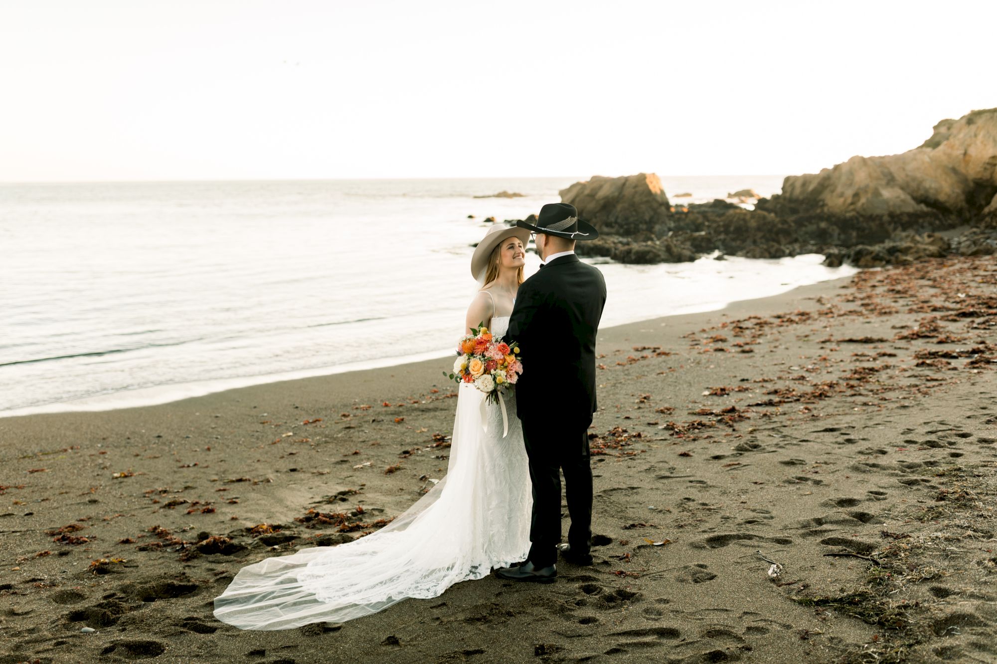 A bride and groom in wedding attire stand on a sandy ocean beach, holding a bouquet with waves and cliffs behind them.