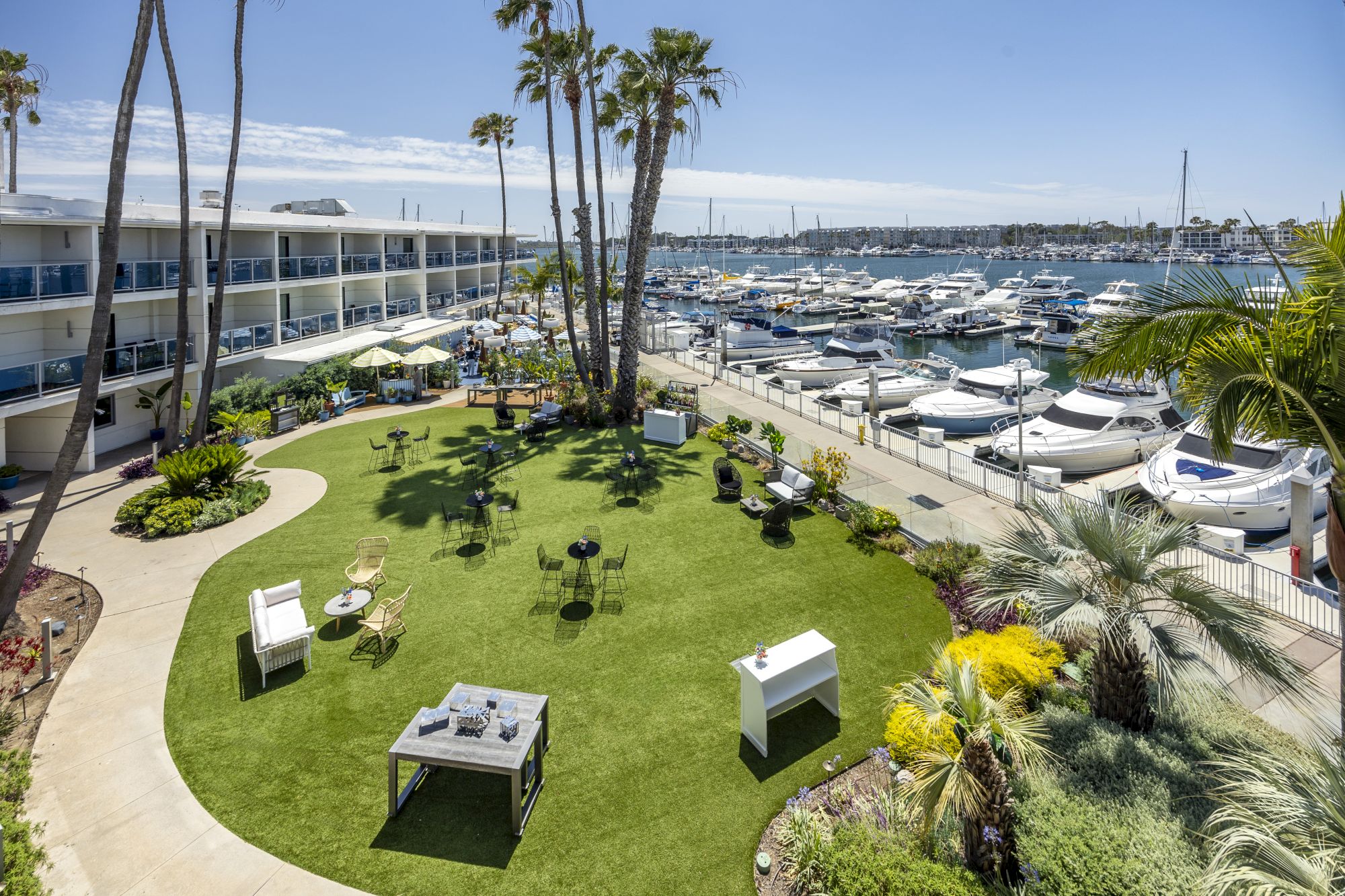 Tropical hotel courtyard with palm trees, manicured lawn, outdoor dining, and a marina packed with sailboats along the coast.