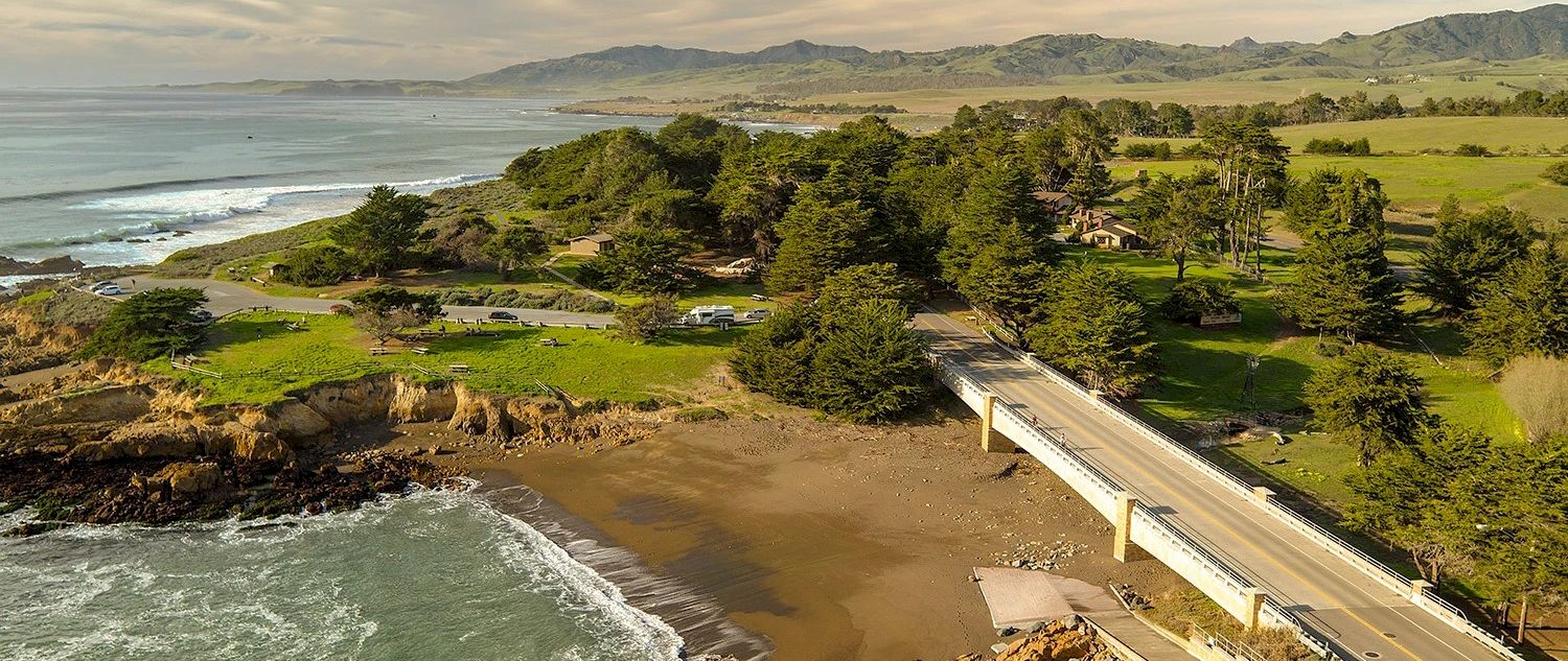 A coastal view with a long bridge road over a sandy beach, waves rolling in, and green hills and mountains in the distance.