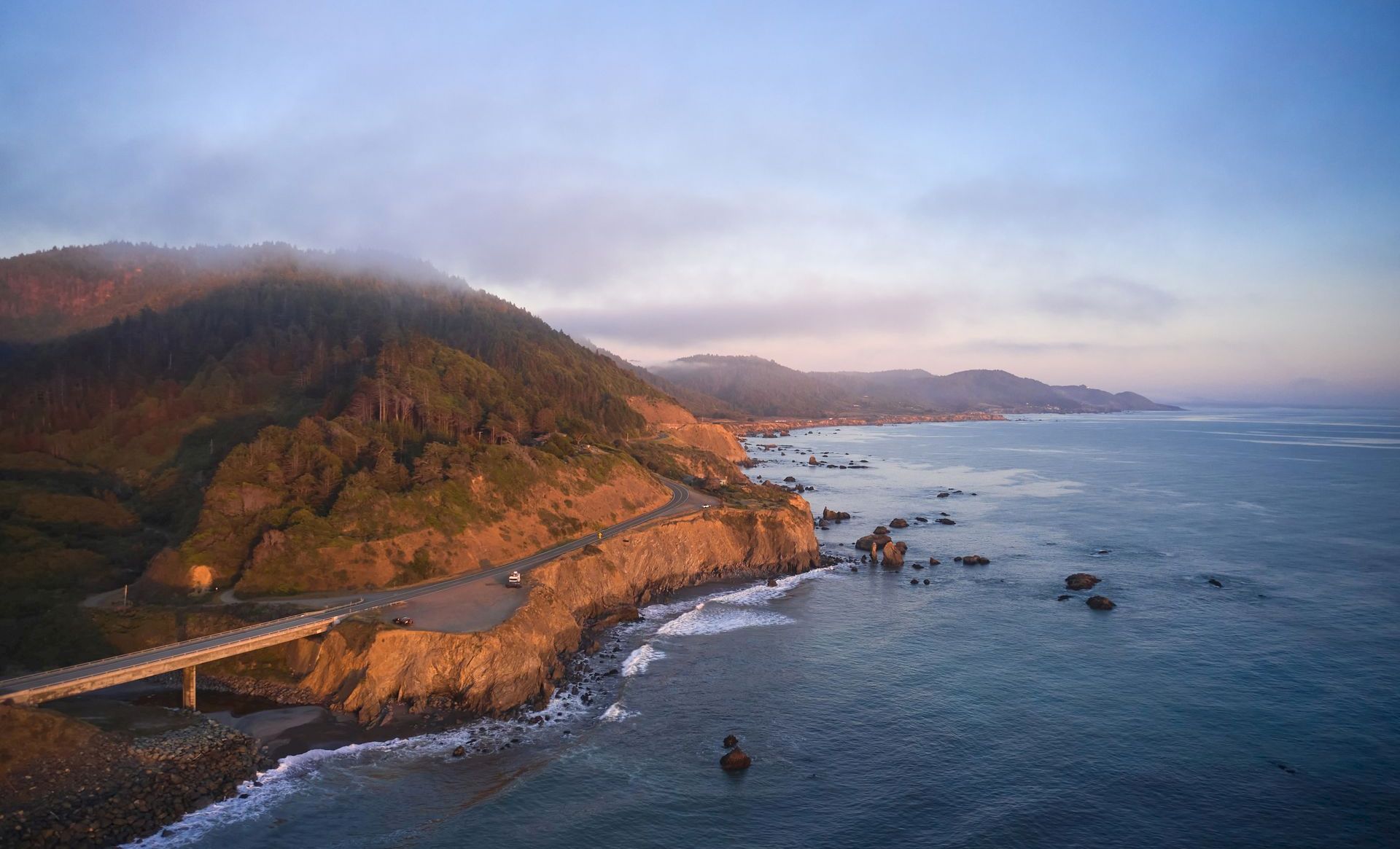 A winding coastal highway hugs cliffs beside a rocky beach, with waves rolling in under a misty, colorful sky.