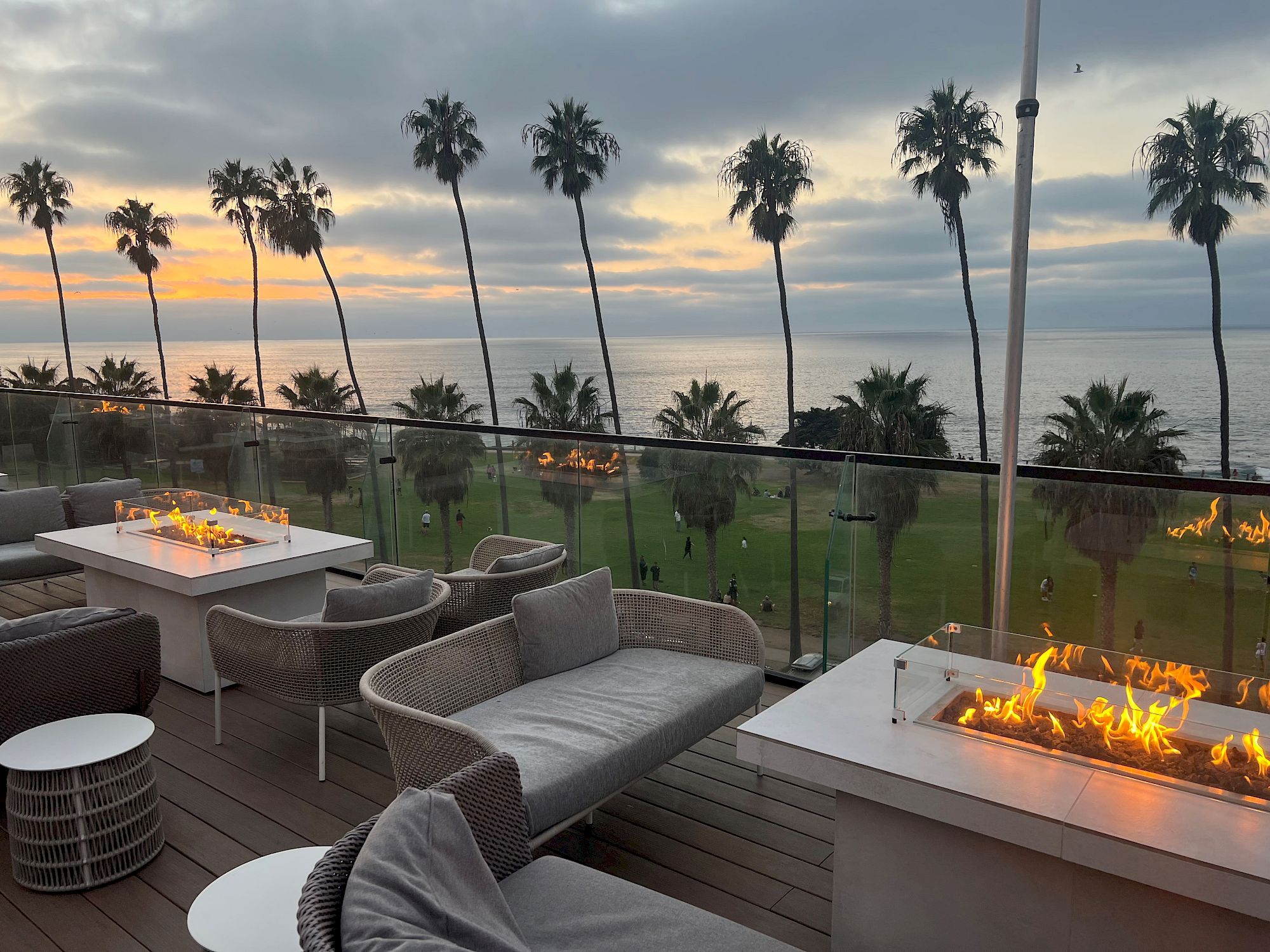 An ocean sunset with tall palm trees, plus a beachfront patio featuring glowing fire pits and outdoor seating by the glass railing.