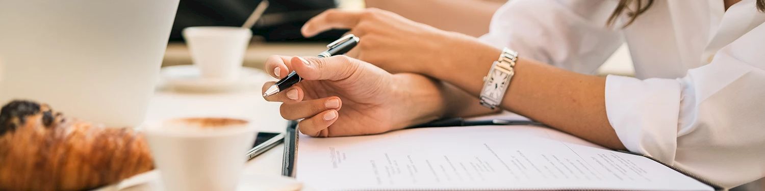 A person in white sleeves is writing/signing paperwork with a pen at a desk, suggesting a meeting or contract review.