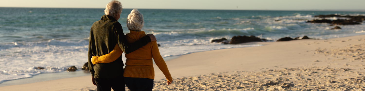 An elderly couple strolls along a sandy beach, watching waves roll in at sunset.
