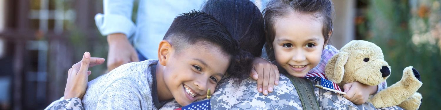 Two children smile and hug, holding an American flag-themed toy and a plush teddy bear in an outdoor setting.