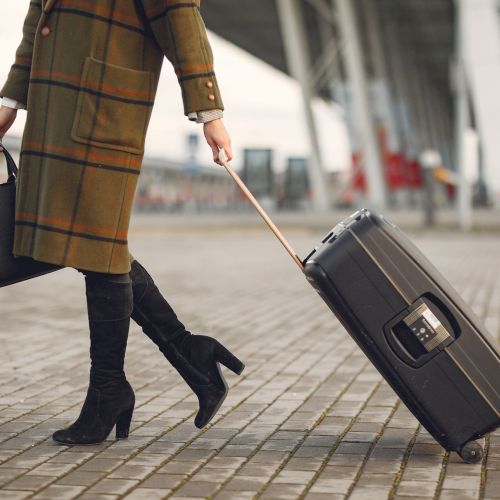 A person in a plaid coat walks at a station or airport, pulling a rolling suitcase, carrying a tote bag and boarding card.