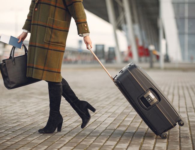 A person in a plaid coat walks at a station or airport, pulling a rolling suitcase, carrying a tote bag and boarding card.