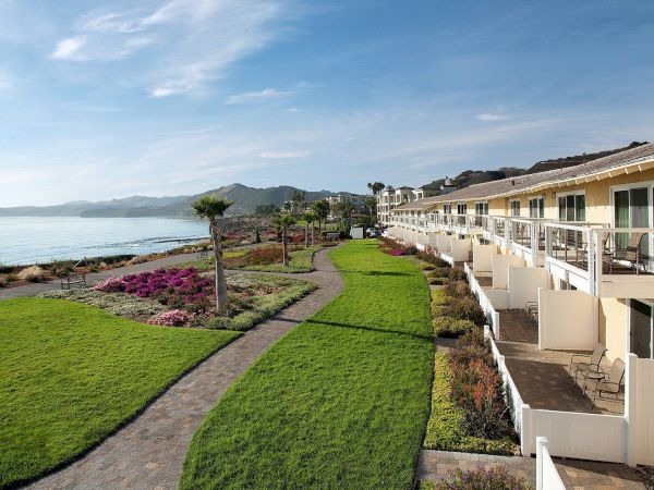 A seaside resort with white balconies, a manicured green garden and colorful flowers beside the ocean under a blue sky.