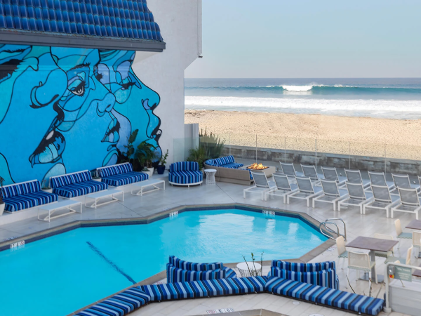 A seaside hotel patio with a blue pool, striped loungers, white chairs, and a big blue graffiti mural beside the ocean.