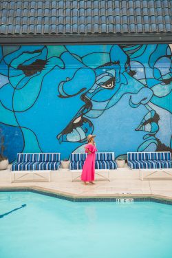A woman in a bright pink dress stands by a pool beneath a large blue graffiti mural of a cartoon character.