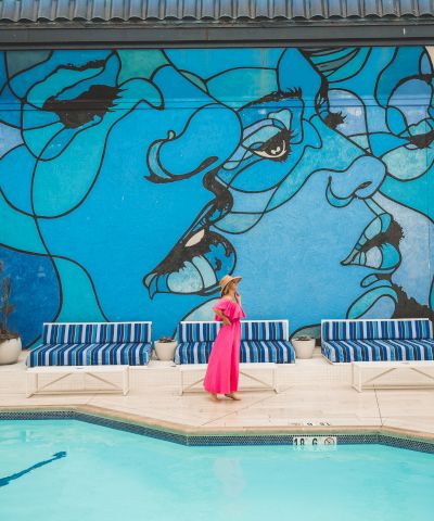 A woman in a bright pink dress stands by a pool beneath a large blue graffiti mural of a cartoon character.