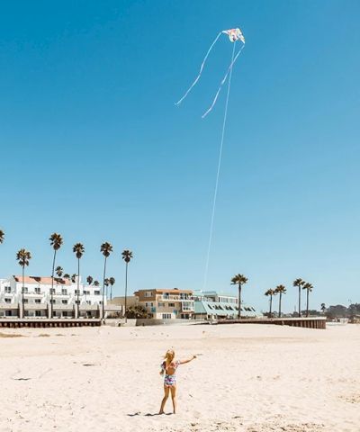 A child plays on a sunny sandy beach as a kite flies high against a blue sky.