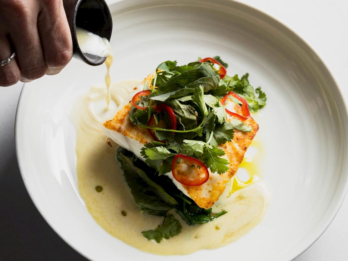 A chef drizzles sauce over a plated piece of fish topped with greens and sliced peppers.