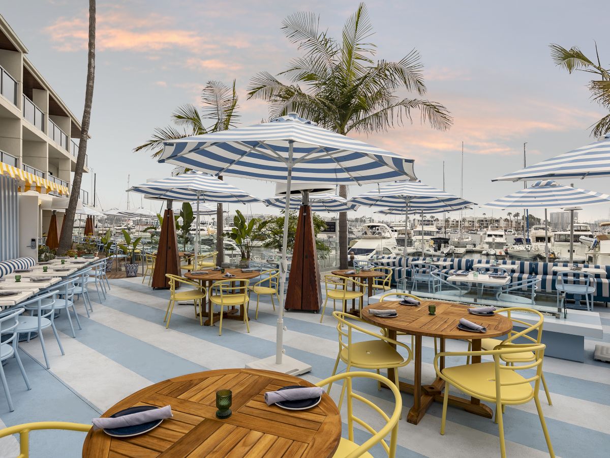 A marina-side patio with striped umbrellas, wooden tables, palm trees, and docked boats under a sunset sky.