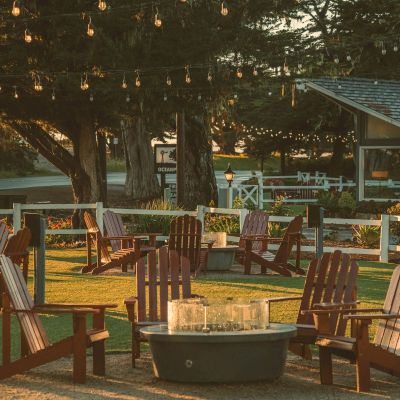 An outdoor patio at sunset with wooden chairs, a stone fire pit, warm string lights, and trees around a grassy lawn.