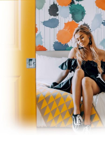 A woman sits on a patterned bed in a colorful room, smiling as she looks down in front of a bright mural wall.
