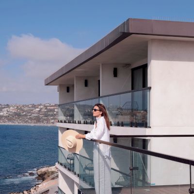 A woman in a white outfit stands on a seaside terrace atop a modern cruise ship, with blue ocean and a coastal town behind her.