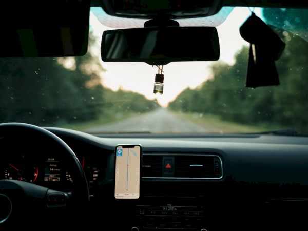 Inside a car, a phone GPS and rearview mirror show a drive down a sunlit road with greenery on both sides.