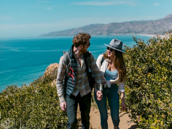 A hiking couple strolls along a sandy coastal trail with backpacks, looking out over the blue ocean and distant hills.
