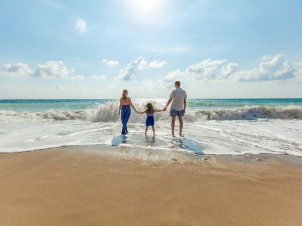 A family of three walks holding hands in the surf at a sunny beach, with waves splashing behind them.