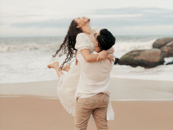 A couple embraces on a sandy beach at the ocean, with the woman lifted and waves behind them.