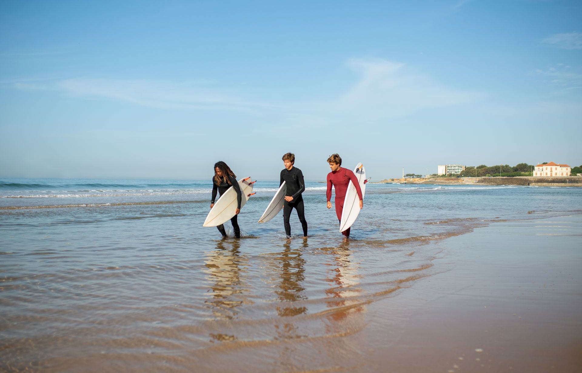 Three surfers walk in shallow water on a beach, carrying white boards under a blue sky.