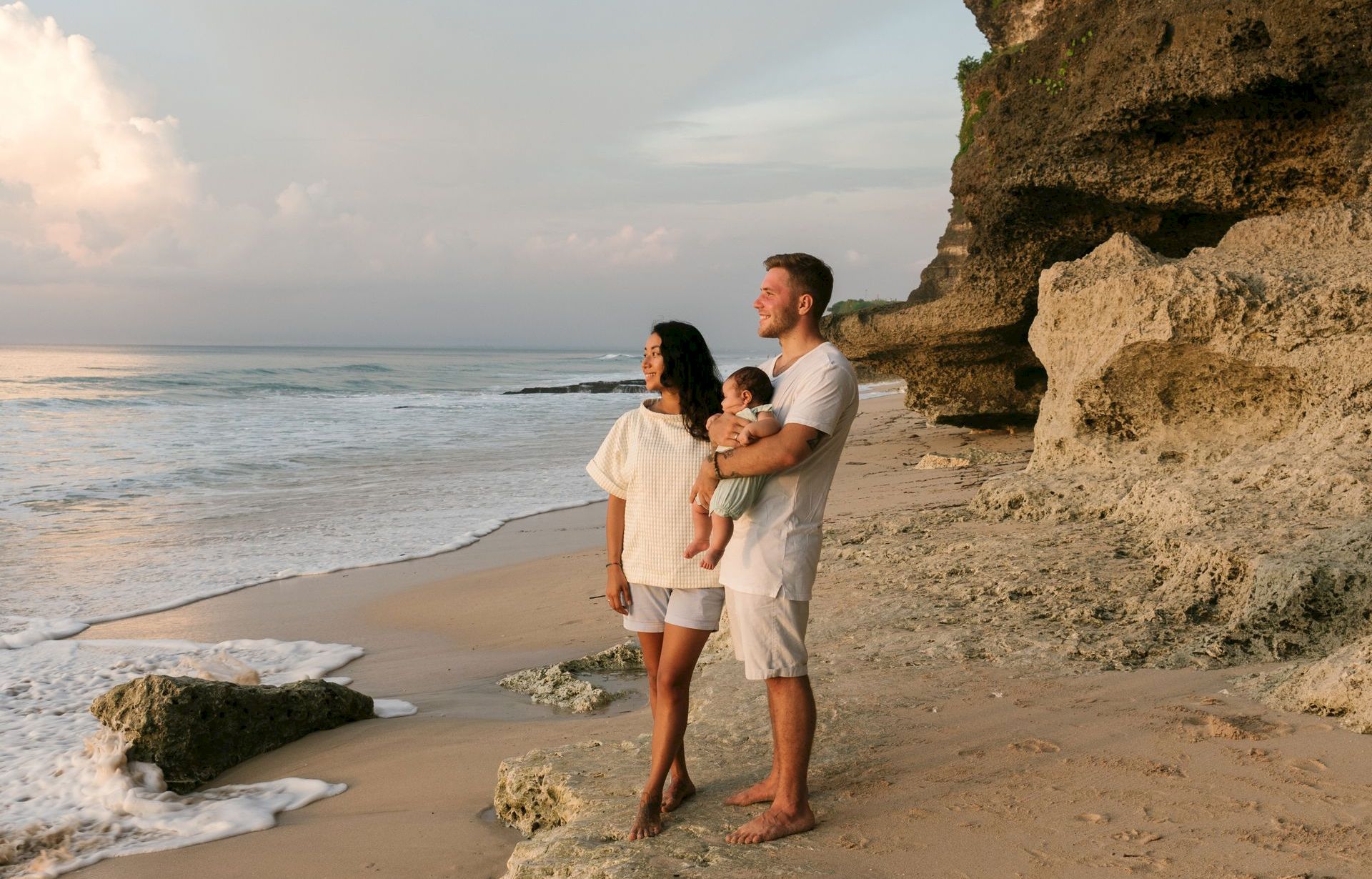 A family of three&mdash;parents holding their baby&mdash;stands on a sandy beach by ocean waves and cliffs at sunset.