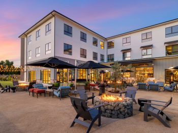 A modern courtyard patio at dusk with a stone fire pit, outdoor seating, black umbrellas, and a white multi-story building glowing with lights.