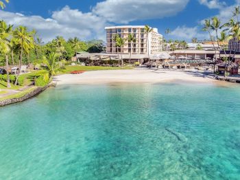 Tropical resort view with palm trees, a turquoise lagoon, and a beachfront hotel terrace with umbrellas and loungers.