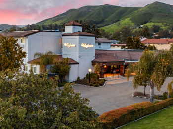 A quiet hotel with a driveway, palm trees, and green hills in the background at dusk, with &ldquo;Wayfare&rdquo; on the entrance.