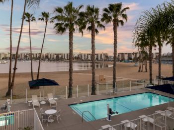 A sunny resort scene with tall palm trees, a turquoise swimming pool, and a sunset harbor view with boats and city buildings.