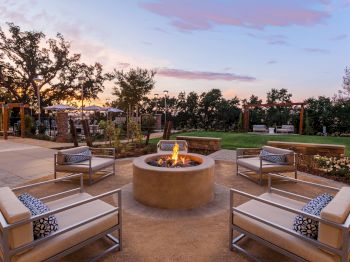 An outdoor patio at sunset with a round fire pit, cushioned lounge chairs, landscaped greenery, and warm string lights.