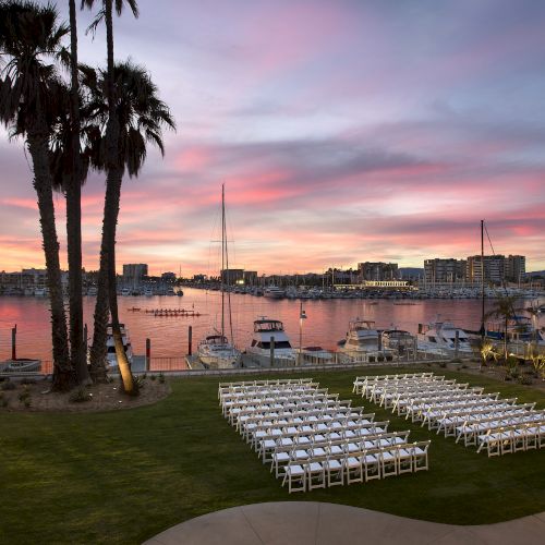 A waterfront venue at sunset with palm trees, rows of white chairs, and docked sailboats.