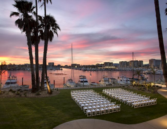 A waterfront venue at sunset with palm trees, rows of white chairs, and docked sailboats.