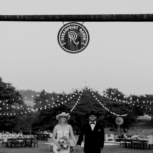 Black-and-white wedding photo: a couple stands beneath a &ldquo;Clean Point Ranch&rdquo; sign with string lights and chairs in the background.