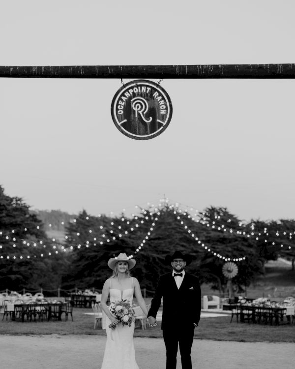 Black-and-white wedding photo: a couple stands beneath a &ldquo;Clean Point Ranch&rdquo; sign with string lights and chairs in the background.