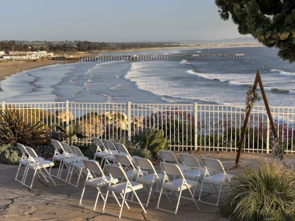 A sunny beach scene with rolling waves, a long pier, and lounge chairs on a terrace overlooking the shoreline.