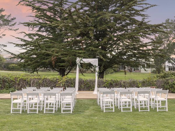 Outdoor wedding ceremony setup with rows of white chairs and a floral aisle arch beneath a huge green tree at sunset.