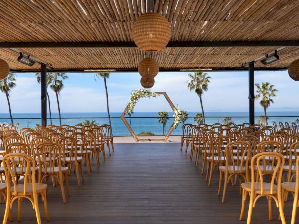 Outdoor beachfront wedding setup with rows of chairs, floral arch, hanging lanterns, and ocean views.