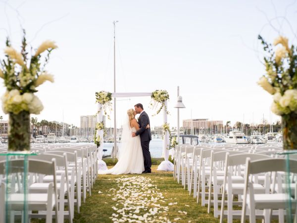 A bride and groom kiss at an outdoor waterfront wedding ceremony under a floral arch, with white chairs and sailboats in view.