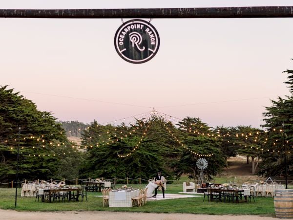An outdoor event setup with tables and string lights in a forest garden, topped by a &ldquo;Greenpoint Ranch&rdquo; logo sign.