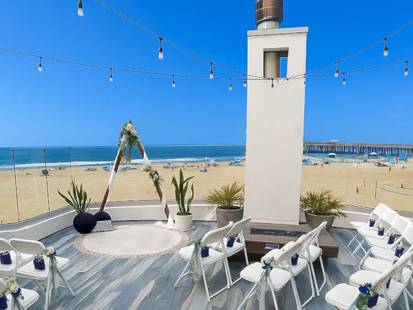 A beachfront event setup with white chairs and blue accents, plus a tall smoking-style column and string lights under a clear blue sky.