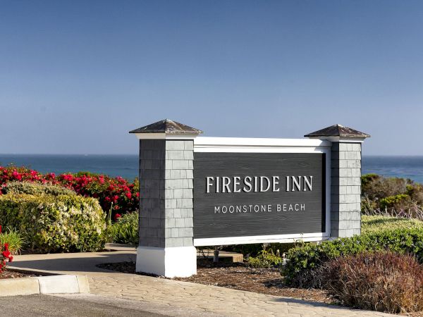 A Fireside Inn sign for Moonstone Beach beside colorful gardens and the ocean under a clear blue sky.