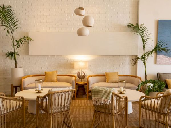 A bright patio lounge with beige sofas, wicker chairs, hanging lamps, and tall palm plants by a brick wall.