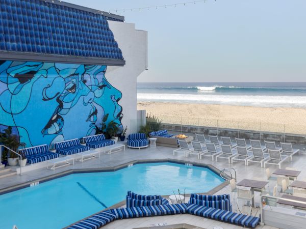 A seaside patio with a blue pool, striped loungers, a large mural on the wall, and ocean waves beyond the terrace.