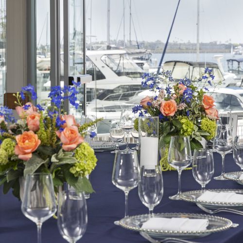 A waterfront banquet table set with colorful flowers, glassware, and silverware, overlooking moored yachts through large windows.