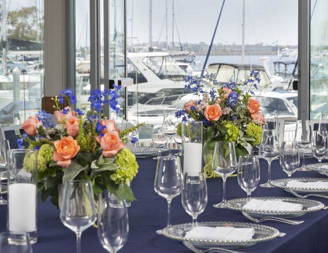 A waterfront banquet table set with colorful flowers, glassware, and silverware, overlooking moored yachts through large windows.