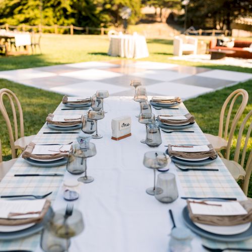 An outdoor reception setup with a long table set for guests&mdash;plates, glassware, menus, and candles in a garden.