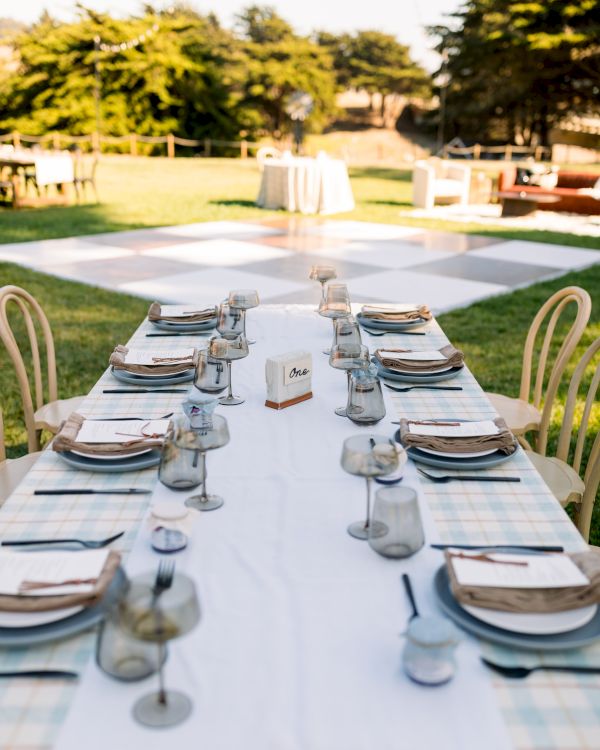 An outdoor reception setup with a long table set for guests&mdash;plates, glassware, menus, and candles in a garden.